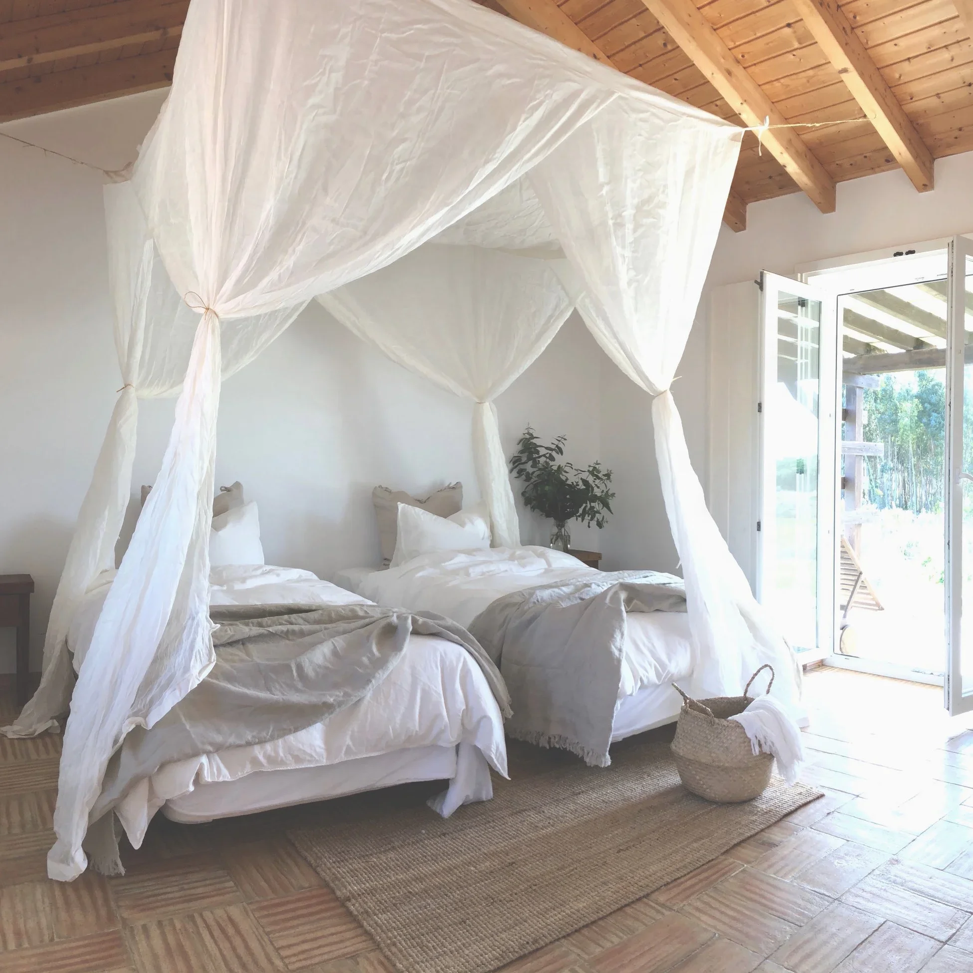 A bedroom with a white bed and a white mosquito net canopy hanging from a wooden ceiling, next to an open door with outdoor view.
