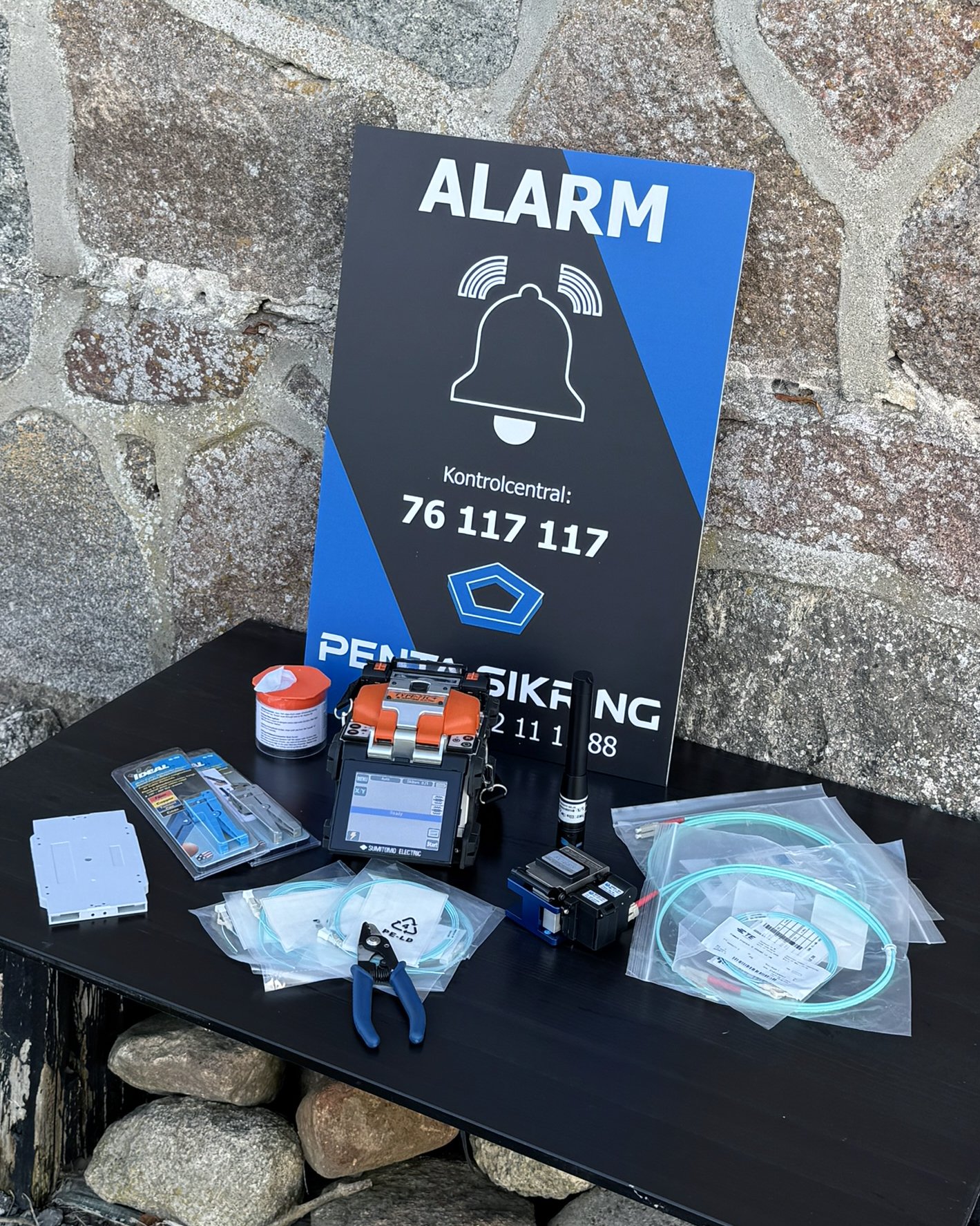 Et sort bord med forskellige værktøjer og elektroniske komponenter, herunder multimeter, ledninger, skruenøgler og en alarmmonitor, sat op foran en stenmur. En plakat med alarm-info og et kontaktnummer står bagpå.