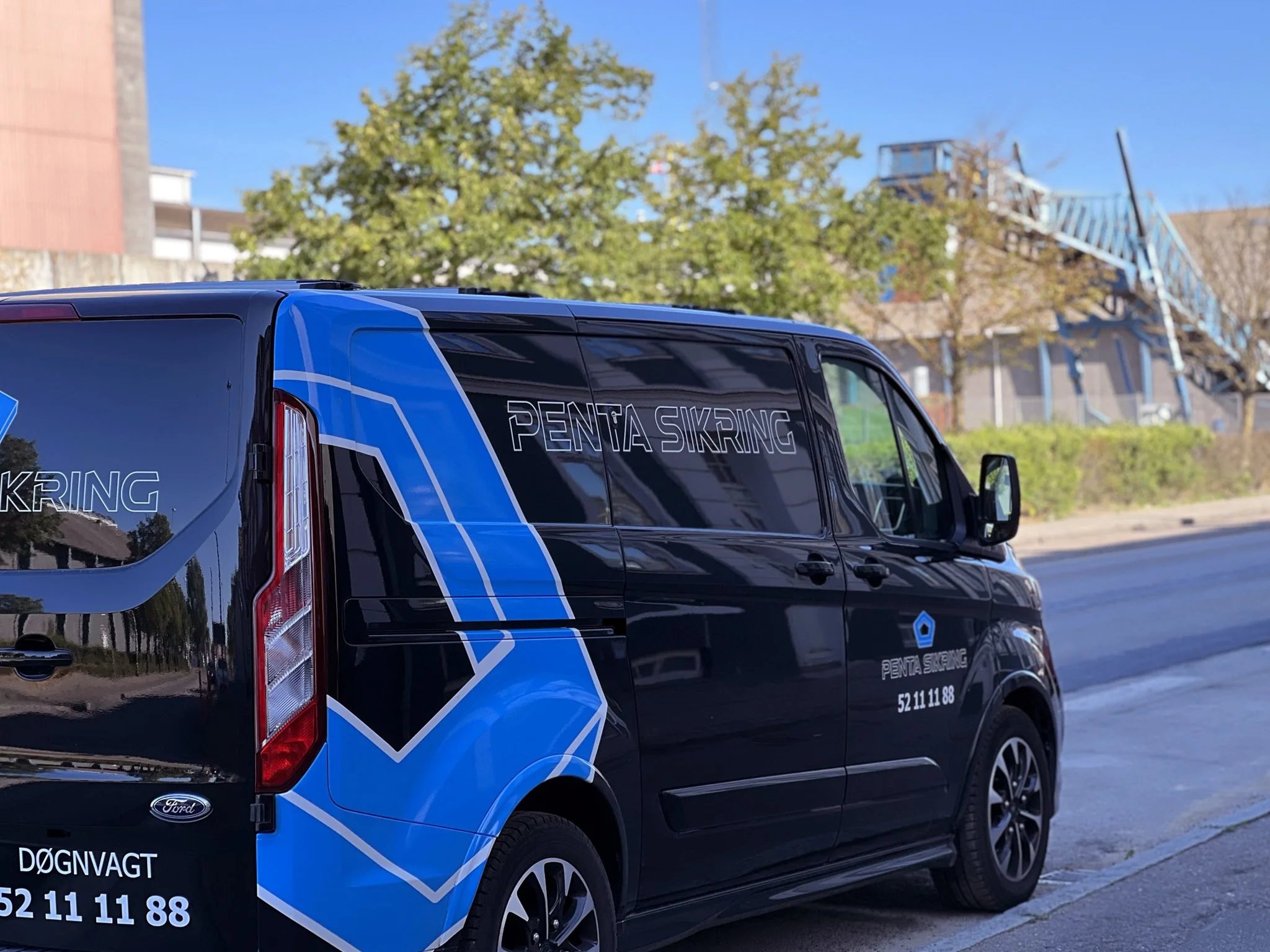 En sort bil med blå og hvide grafik, der har teksten 'Penta Sikring' og kontaktinformation, parkeret ved siden af et træ og en blå trappe i baggrunden.