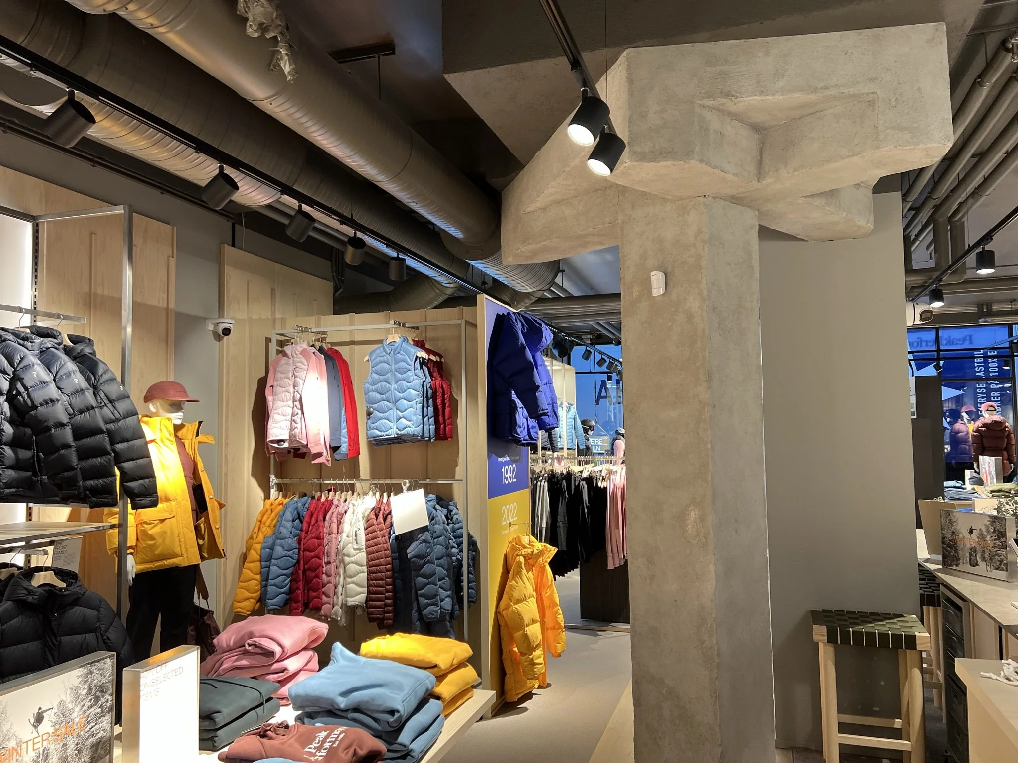 Indretning i en butik med jakker og tøj på væggen og borde, slanke sorte loftlamper og synlige store ventilationsrør i loftet.
På en søjle kan man se en del af alarmssystemet som er sat op af Penta Sikring.