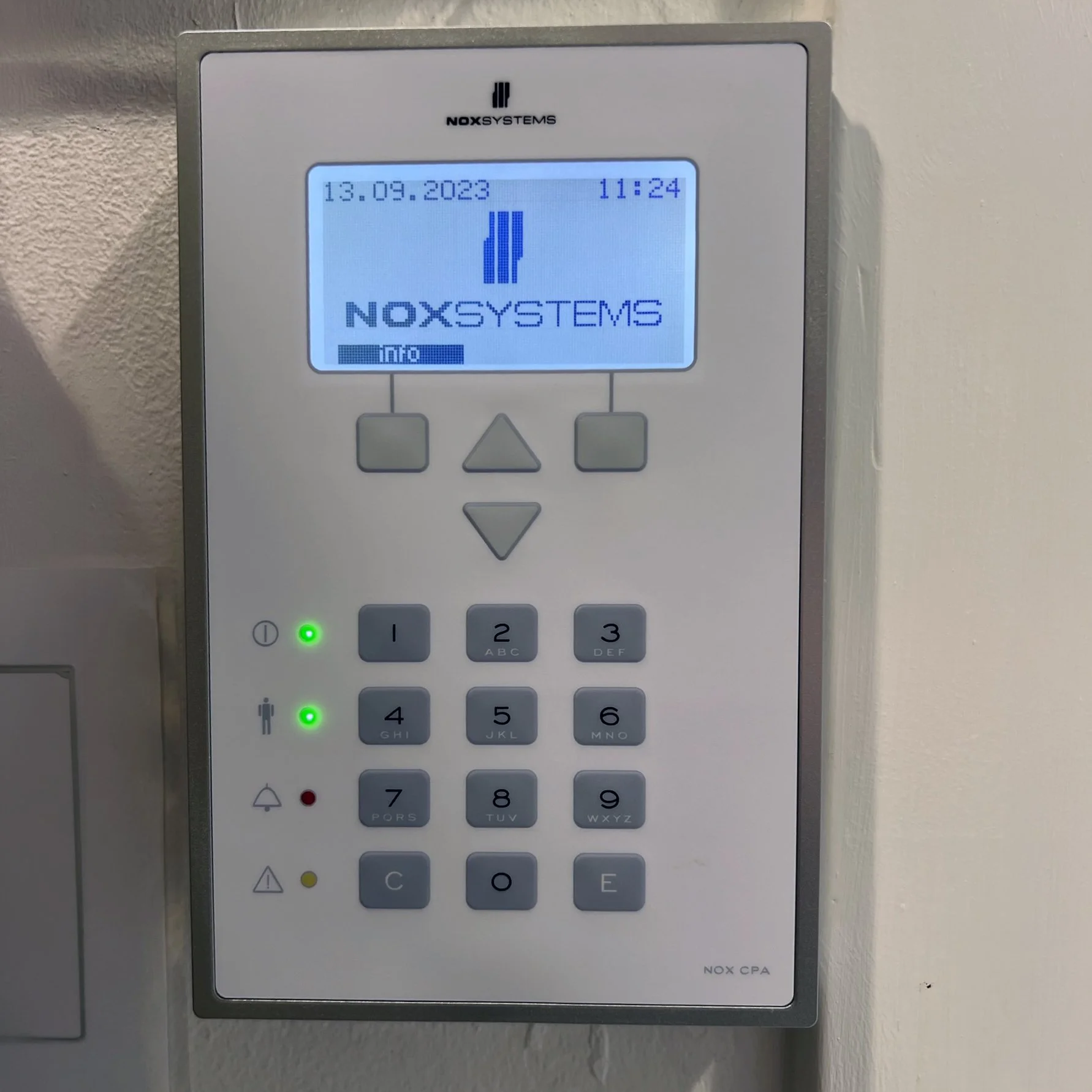 Et adgangskontrolpanel med en skærm, der viser datoen 13.09.2023, klokken 11:24 og teksten 'NOX SYSTEMS'. Der er knapper med tal fra 0 til 9, samt knapper med symboler for reset, alarm, advarsel og adgangskode.