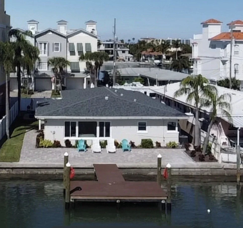 A waterfront house with a dock, small patio, and Adirondack chairs, surrounded by palm trees and neighboring white houses.