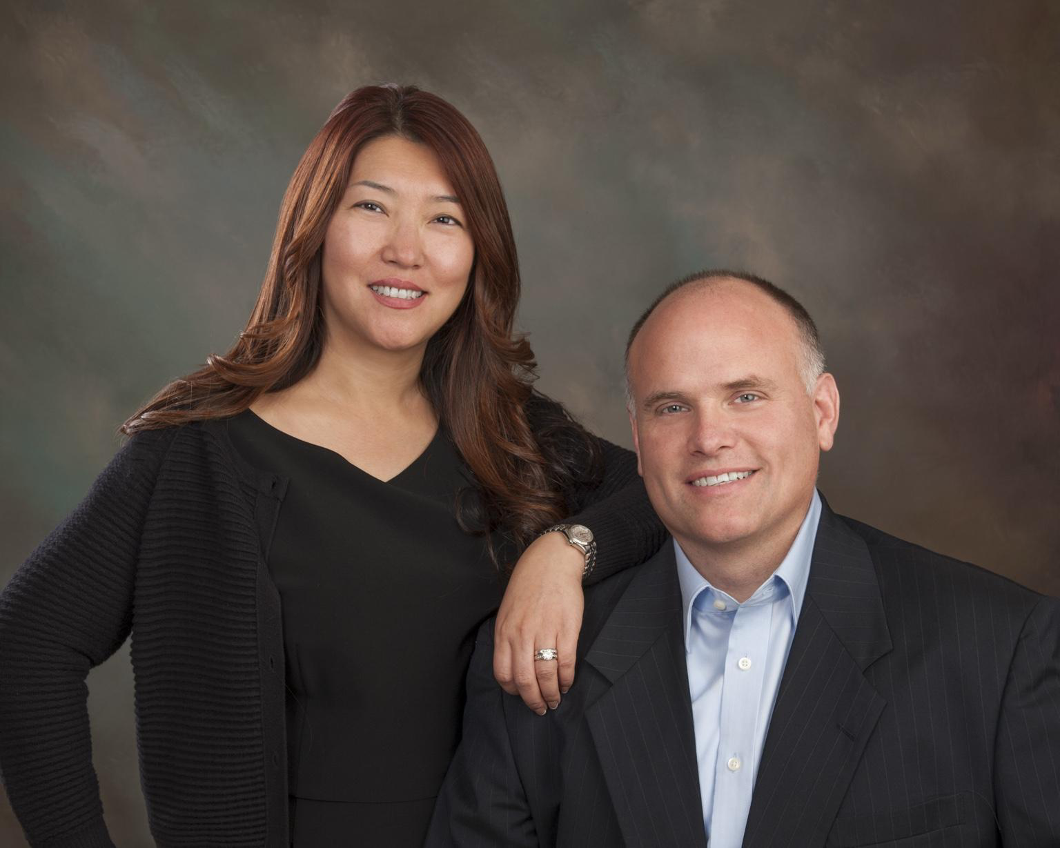 A woman with long, wavy brown hair and a man with a shaved head, both smiling, posing together against a neutral background. The woman is standing with her arm resting on the man's shoulder, and the man is seated in a suit with a light blue shirt.