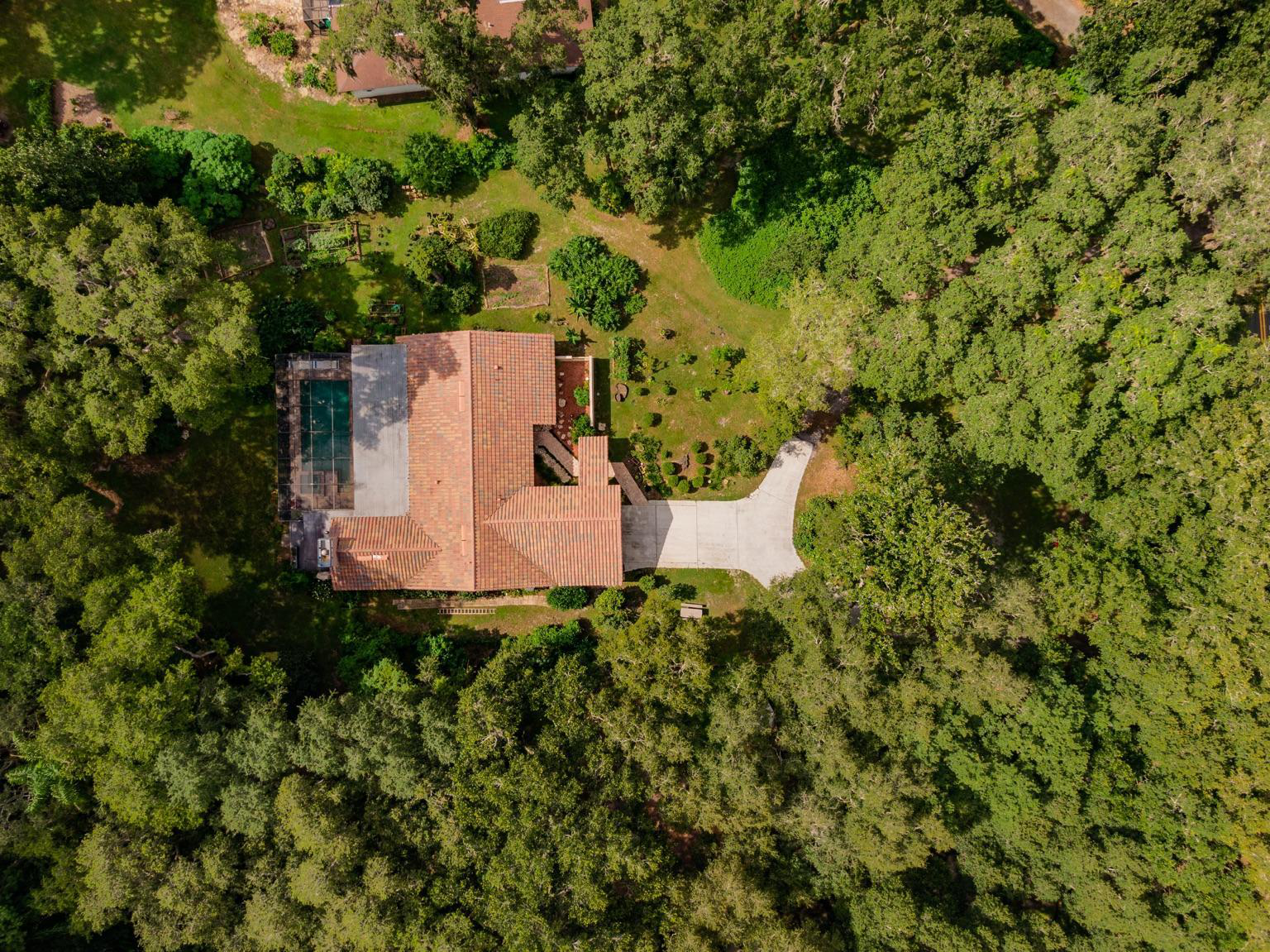 An aerial view of a house with a red-tile roof, surrounded by lush green trees and a spacious backyard. The backyard has a concrete driveway, small garden beds, and an enclosed swimming pool.