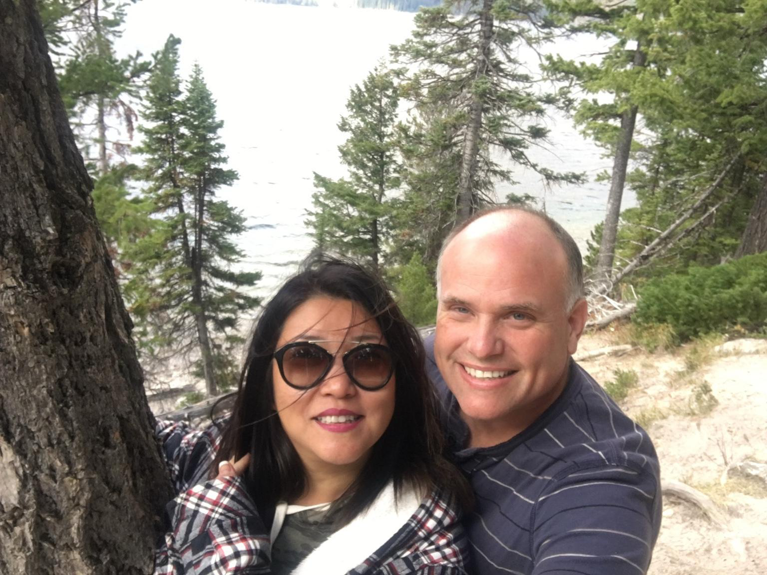 A smiling couple taking a selfie outdoors near a large tree in a forested area by a lake or river.