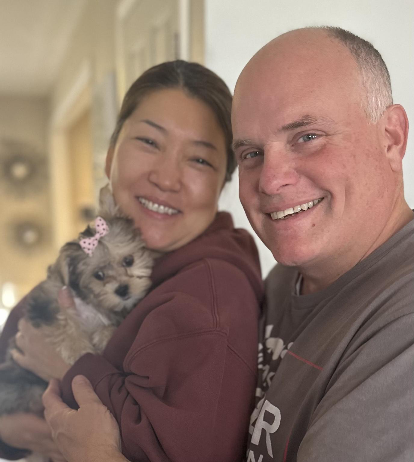 A smiling woman holding a small, gray puppy with a pink bow, standing next to a man with a short haircut, both smiling at the camera.