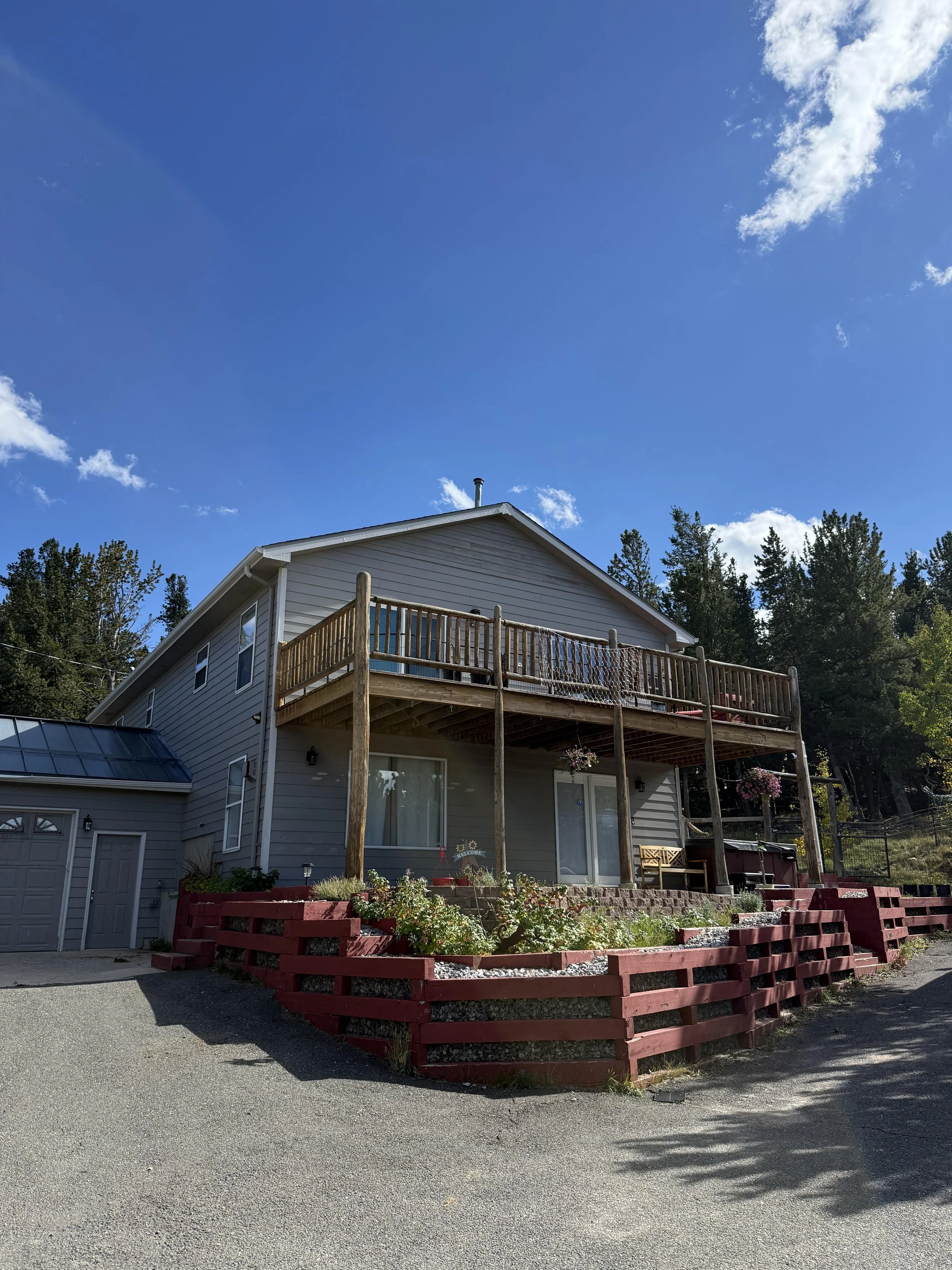 A two-story house with a porch and a balcony, surrounded by trees and a garden with flowers, under a bright blue sky with a few scattered clouds.