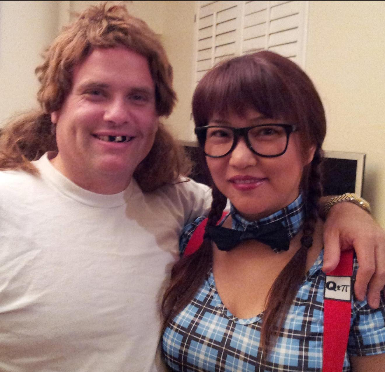 A man with long, curly red hair and missing front teeth smiling with his arm around a woman with glasses, braids, and a checkered shirt. They are indoors near a computer monitor.