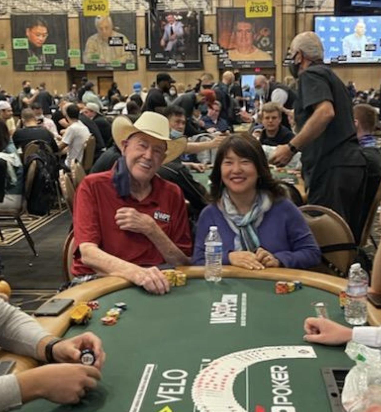Two people sitting at a poker table in a crowded casino or event. The man on the left is wearing a cowboy hat and a red shirt, and the woman on the right is wearing a purple jacket with a scarf. They are smiling and appear to be enjoying their time. In the background, there are many other players, some wearing masks, and large screens displaying images and information.