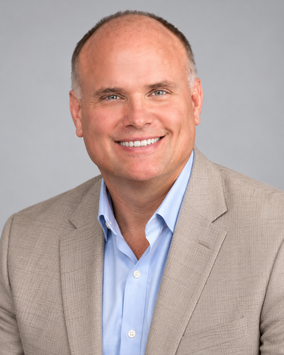 Headshot of a smiling man wearing a beige blazer and light blue dress shirt against a neutral background. Mickey Moore headshot