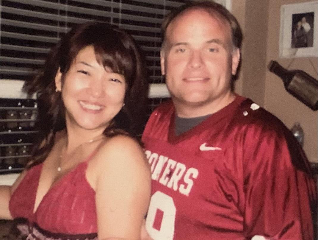 A smiling woman and a smiling man are posing for a photo indoors. The woman has shoulder-length brown hair and is wearing a sleeveless red top. The man has short light brown hair, is wearing a red sports jersey with the word 'SPOKANE' and the number 0 on it, and is standing close to the woman.