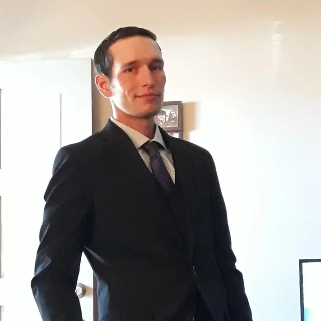 A young man in a black suit, white shirt, and dark tie standing indoors near a plain white wall, with a slight smile.
