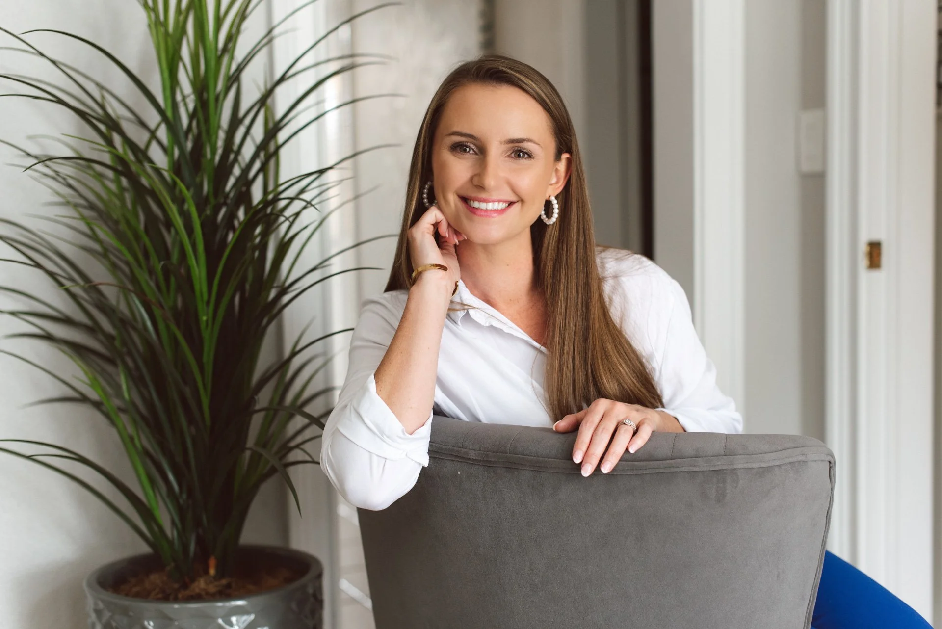 A woman with long brown hair, wearing a white blouse and pearl earrings, smiling and resting her head on her hand while sitting behind a gray chair, with a large potted plant in the foreground and a bright, modern interior background.