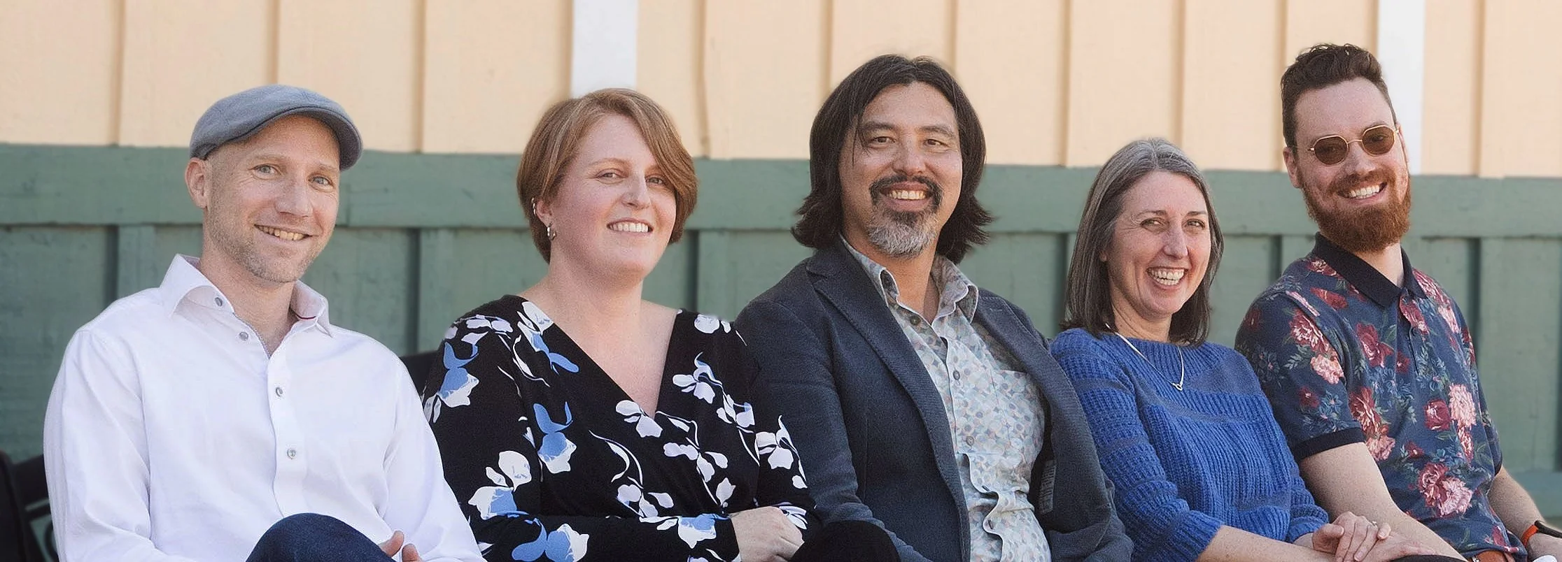 A group of the five employees of Longhouse Architects sitting outdoors in front of a yellow and green wooden fence, smiling at the camera.