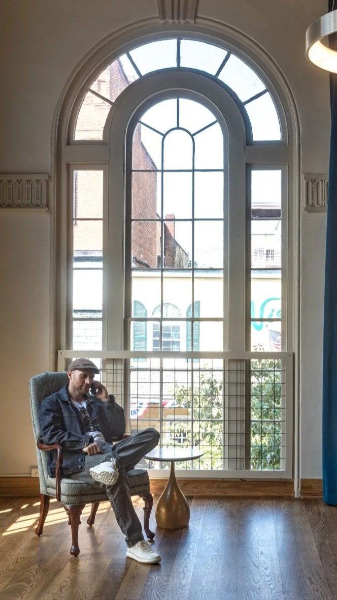 A man sitting in a vintage armchair by a small round table, talking on the phone, in front of a large arched window with multiple panes, in a room with wooden flooring and decorative wall trim.