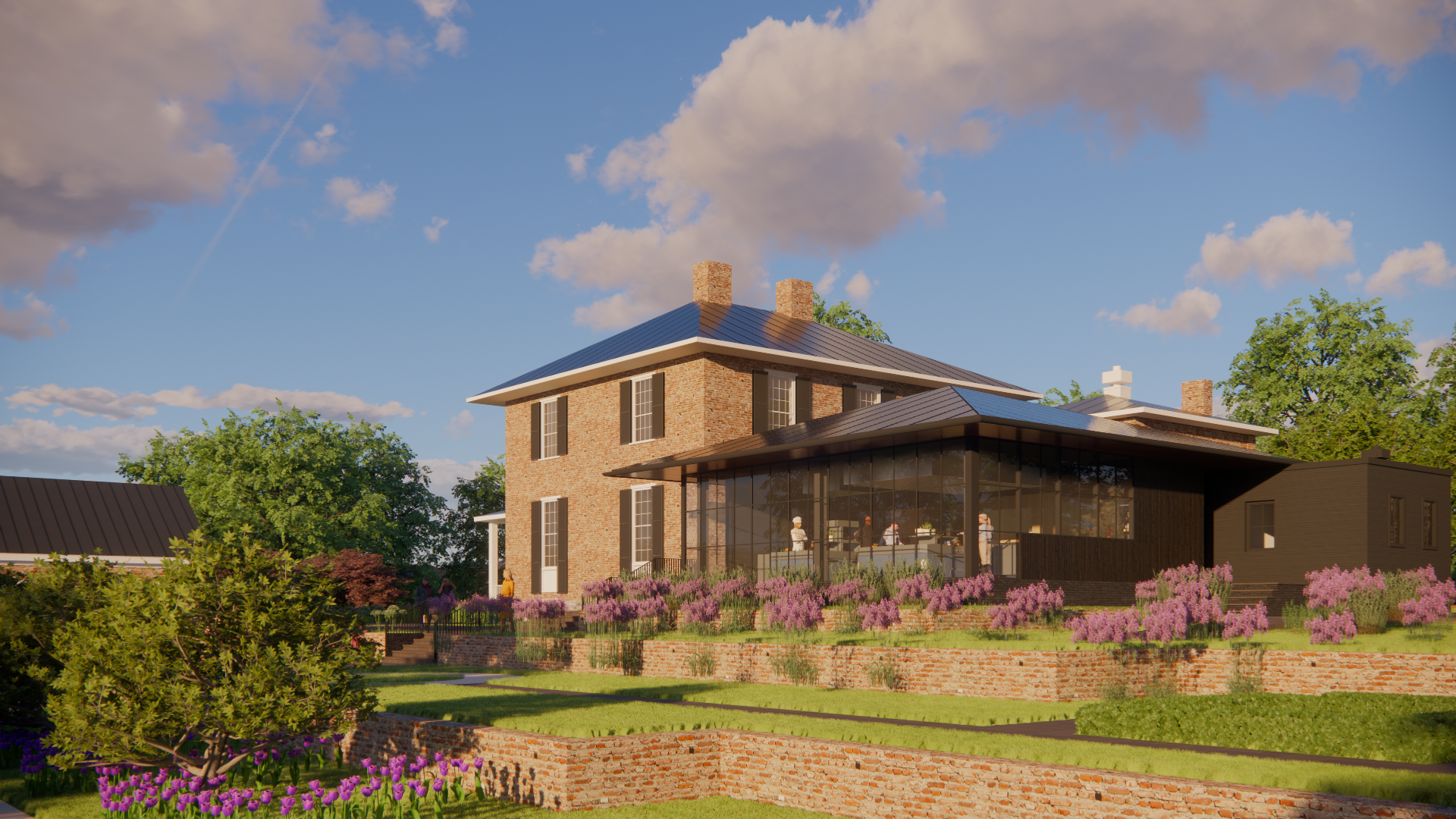A two-story brick manor with a modern glass commercial kitchen addition, surrounded by flowering plants and trees under a partly cloudy sky.