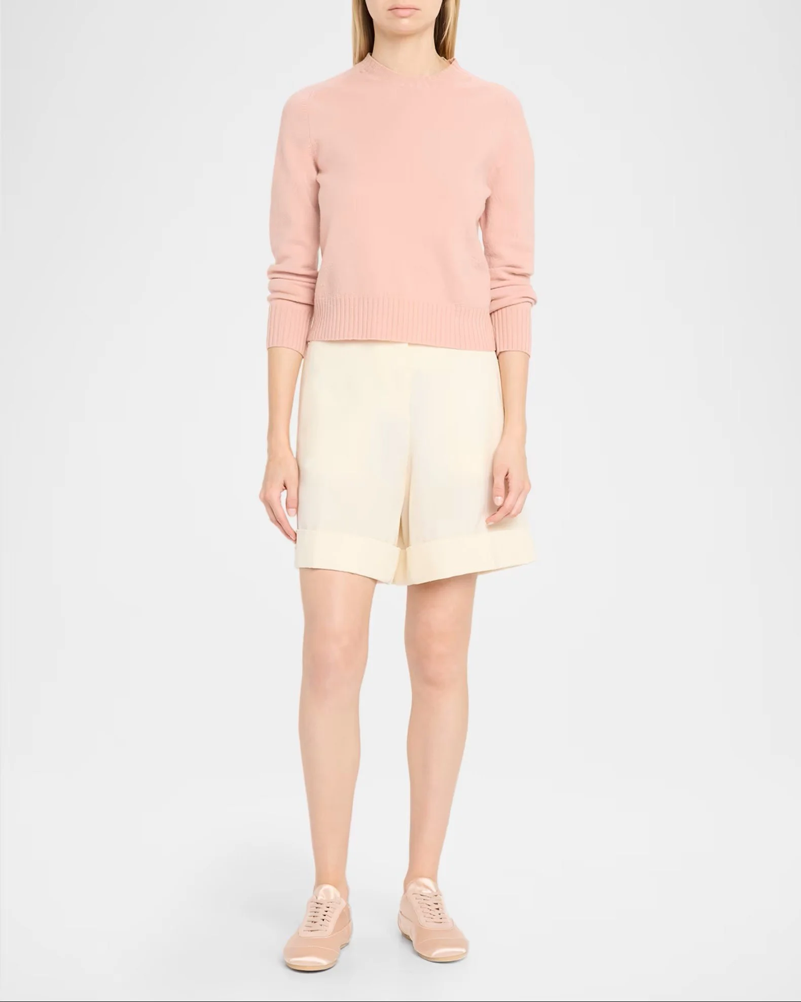 A woman wearing a light pink sweater, cream-colored shorts, and pink sneakers, standing against a plain light background.