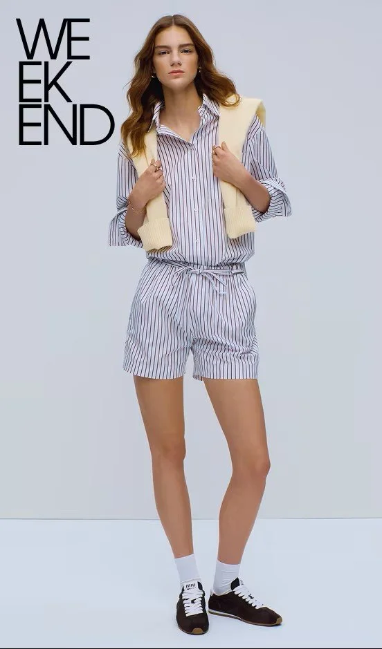 A young woman with wavy brown hair wearing a striped button-up shirt and matching shorts, with a cream vest draped over her shoulders, black sneakers, and white socks, standing against a gray background with the text 'WEEKEND' in black letters.