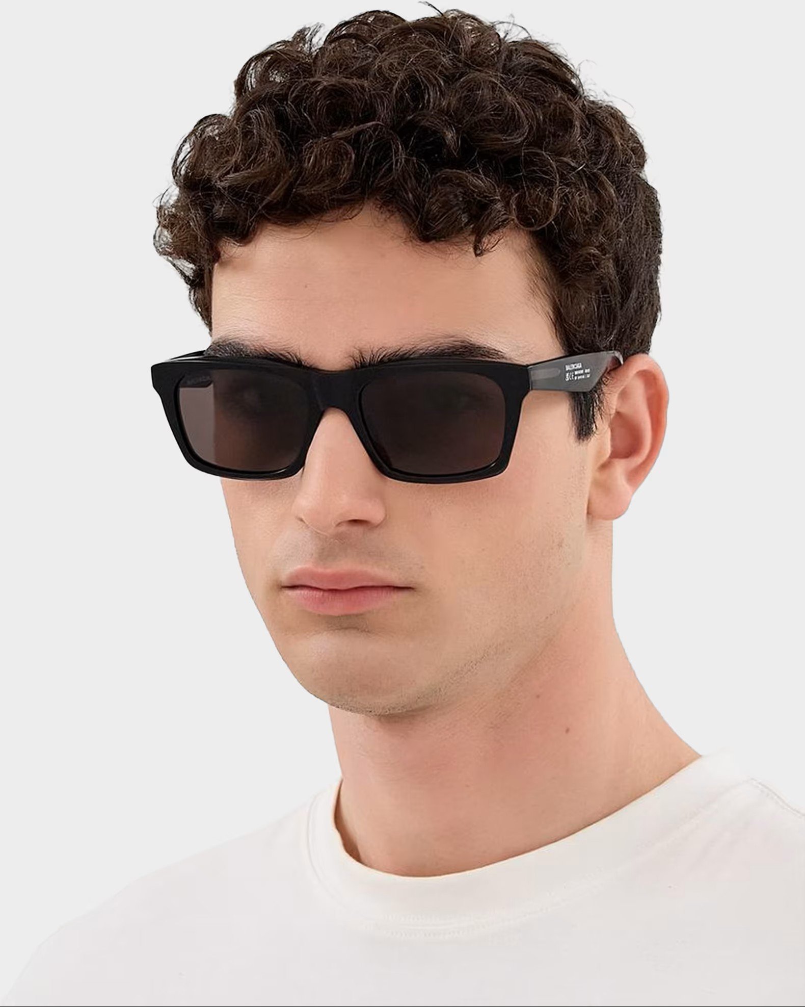 A person with short curly brown hair wearing black sunglasses and a white shirt.