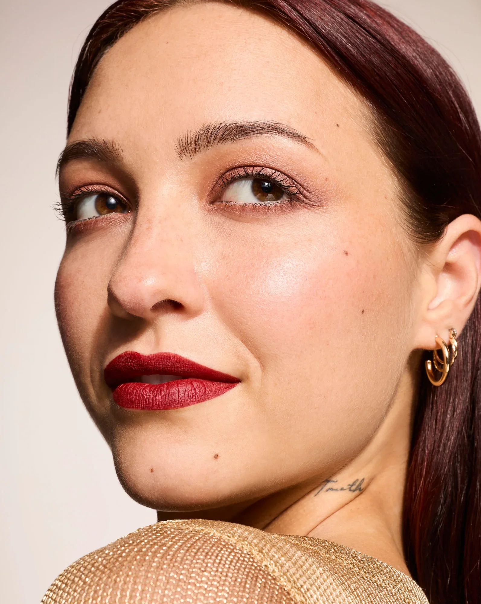 Close-up of a woman's face with red lipstick, minimal makeup, earrings, and a tattoo reading 'Faith' on her neck, wearing a gold textured top.