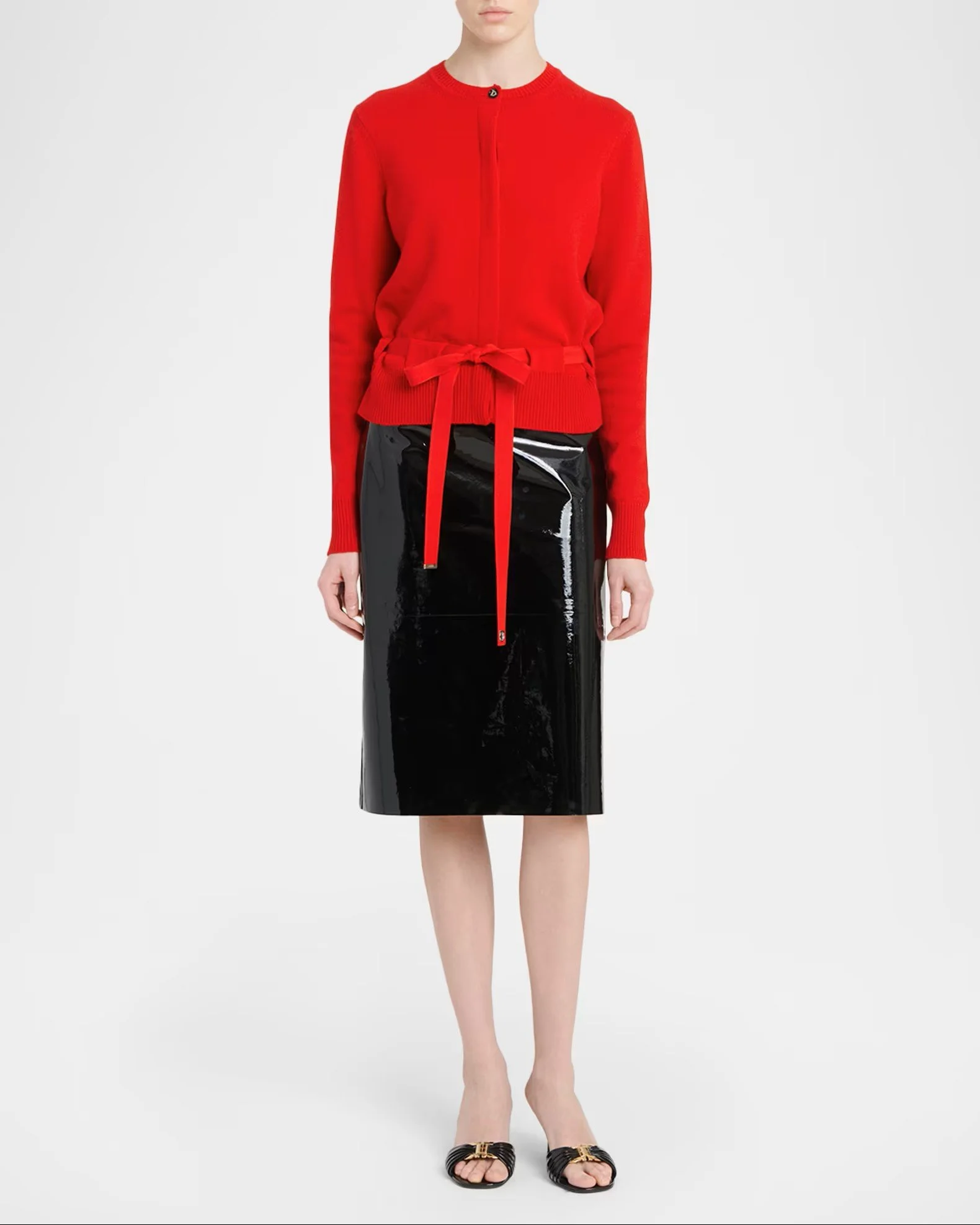 A woman wearing a red zip-up sweater with a bow tie at the waist and a black shiny skirt, paired with black sandals with gold details, standing against a plain white background.