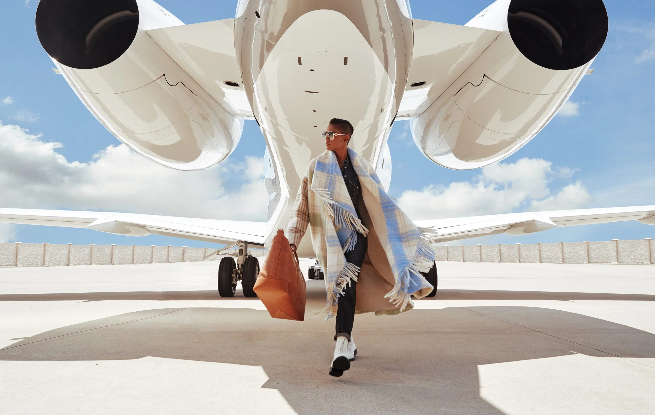 A person walking away from a large private jet aircraft on tarmac, wearing sunglasses, a plaid scarf, and carrying a brown tote bag.