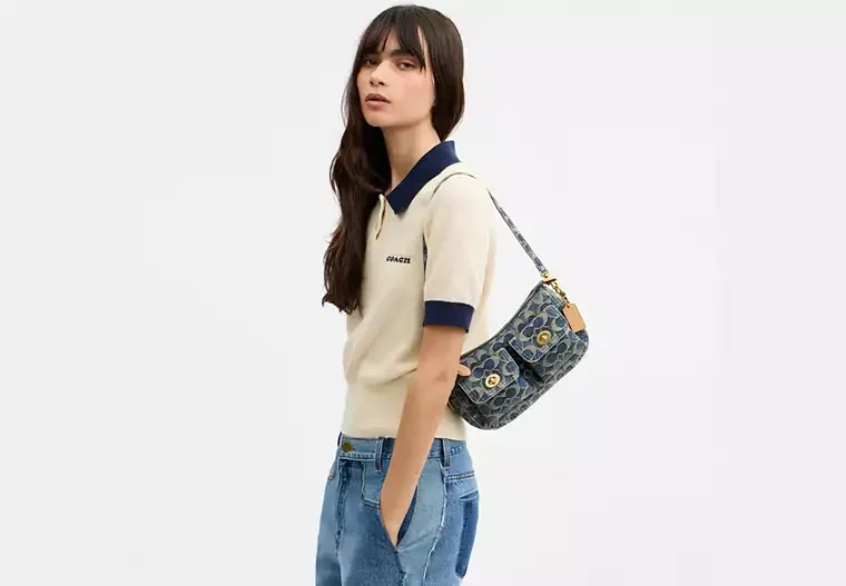 A young woman with long dark hair wearing a beige and navy polo shirt, blue jeans, and carrying a patterned shoulder bag against a plain white background.