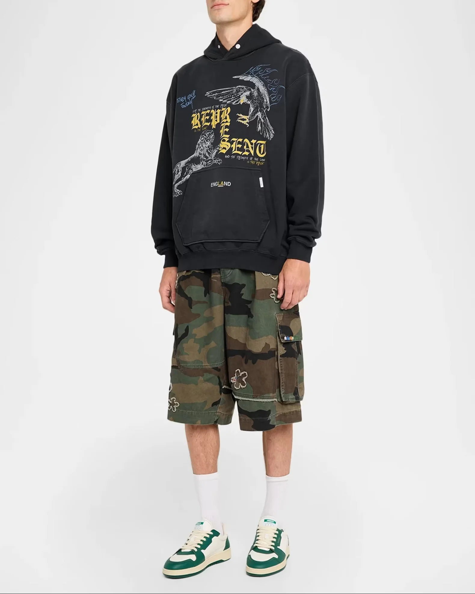 Person wearing a black hoodie with graphic designs and text, camo shorts with patches, white socks, and green and white sneakers, standing against a plain background.