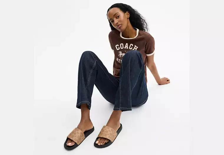 A woman sitting on the floor against a white background, wearing a brown T-shirt with white trim and the word 'COACH' printed on it, dark jeans, and tan slide sandals.