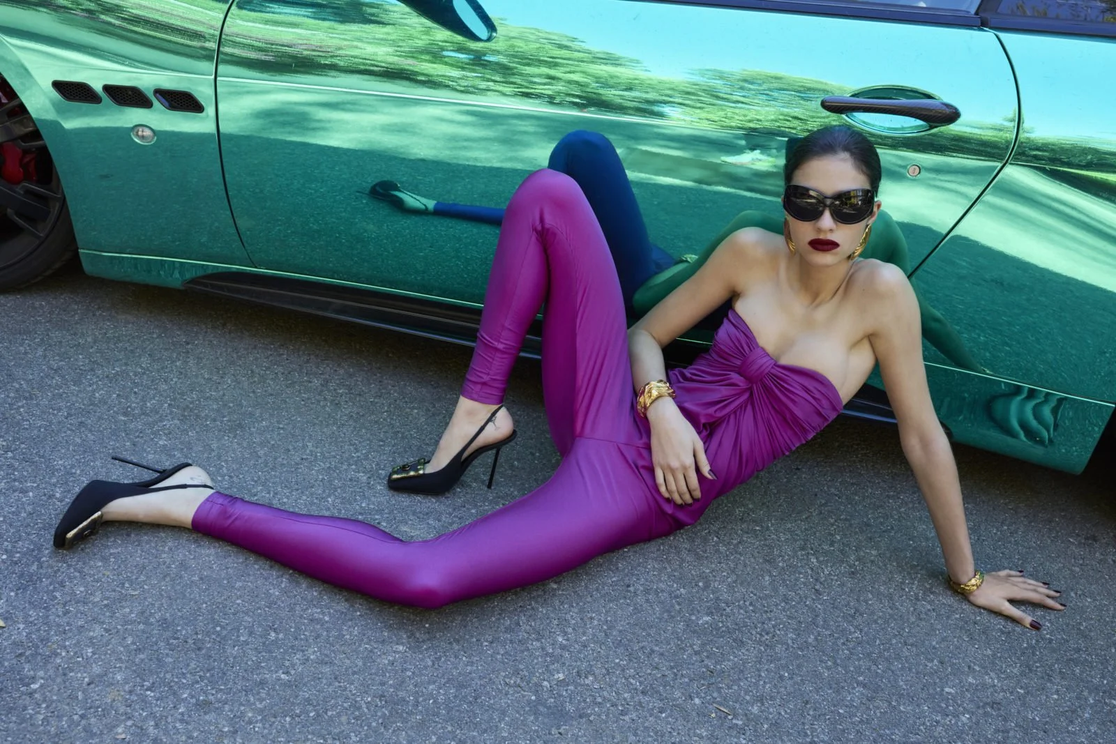 A woman in a purple strapless jumpsuit poses on the ground, leaning against a green sports car. She is wearing black high heels, sunglasses, gold jewelry, and dark lipstick. Her hair is slicked back.