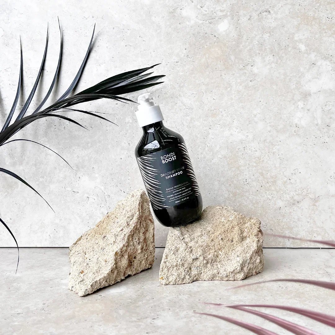 A black bottle of Bondi Boost dandruff shampoo is positioned on two large beige rocks against a light textured background. A black plant with long, curved leaves is partially visible on the left side of the image.