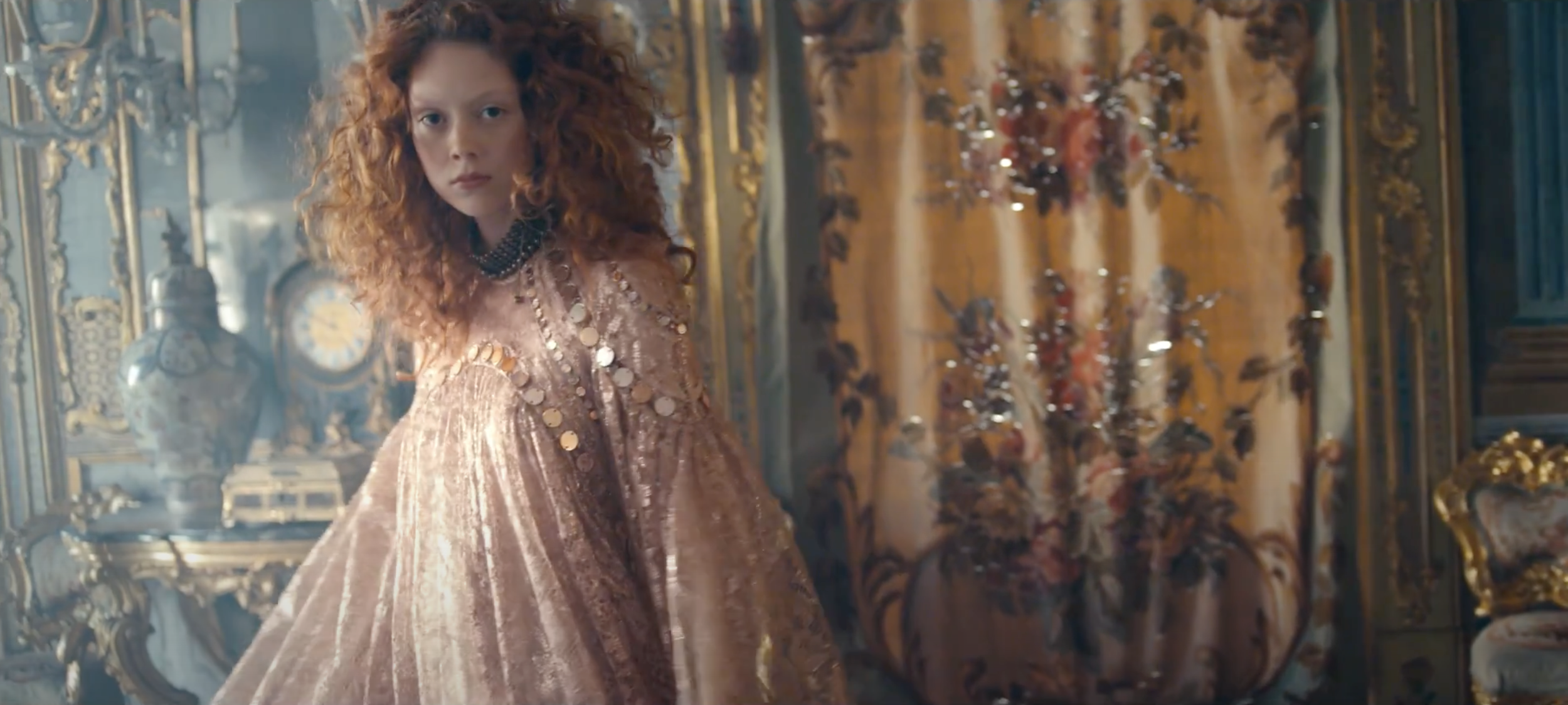 A woman with curly red hair wearing a shiny, pink, sequined gown standing in an ornate, gold-accented room with elaborate wall decorations and vases.