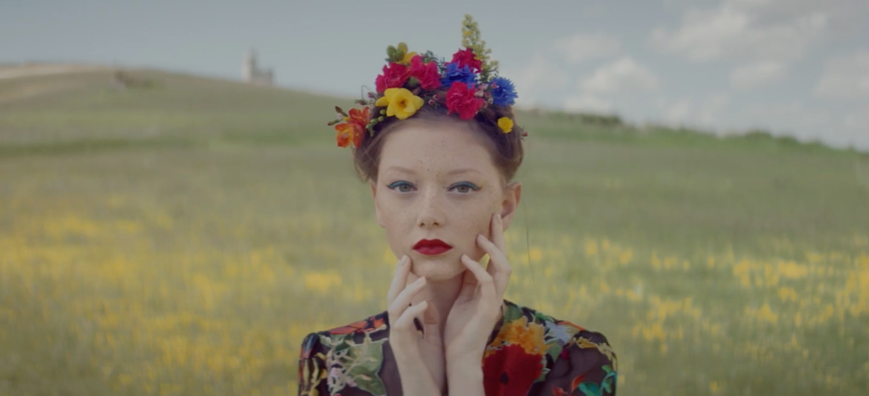 A woman with red hair wearing a colorful floral dress and a crown of flowers on her head, standing in a field of yellow flowers with a clear sky in the background.