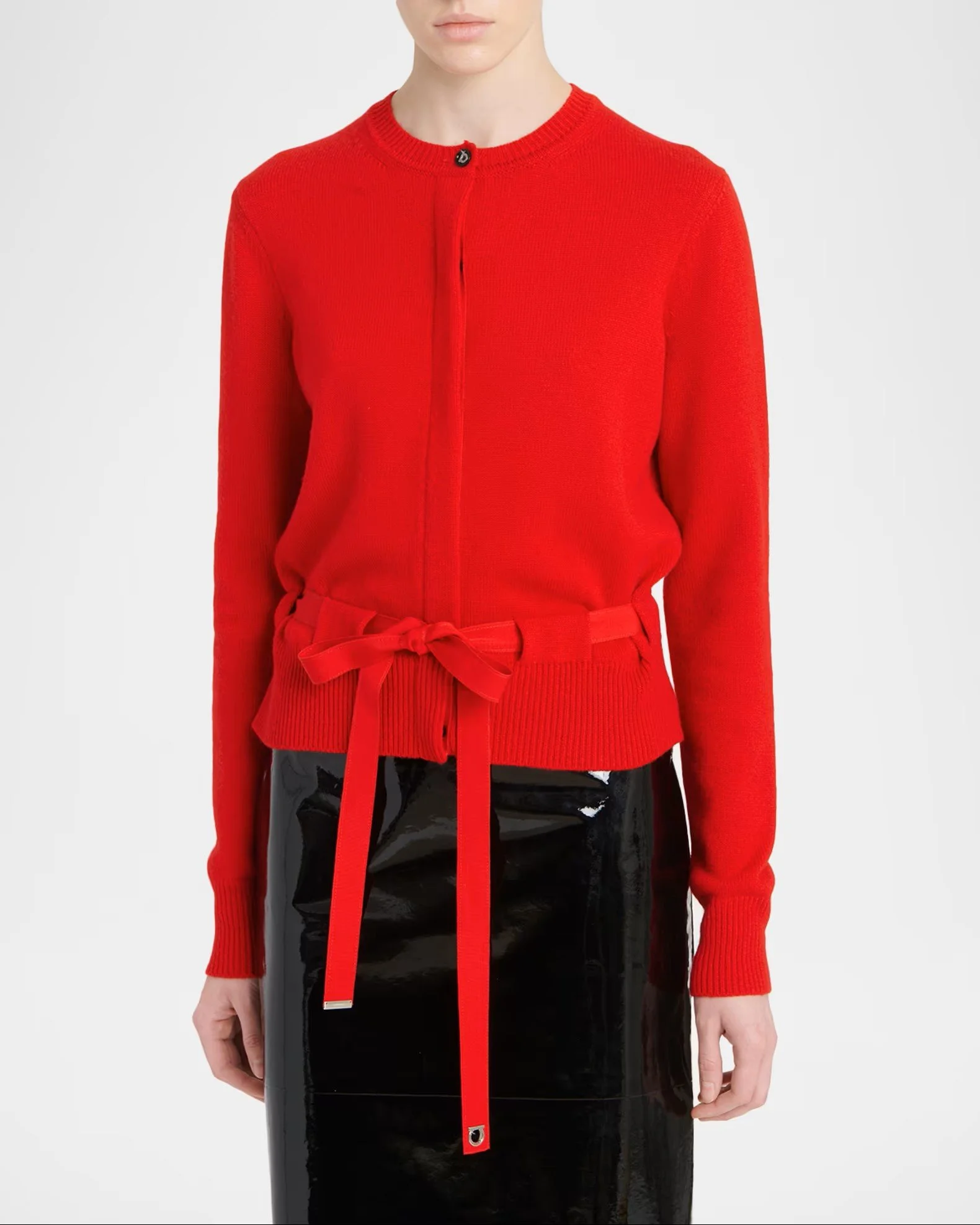 A person wearing a bright red, zip-up cardigan with a matching belt, which is tied in a bow at the waist. The person is also wearing a black, shiny, patent leather skirt.