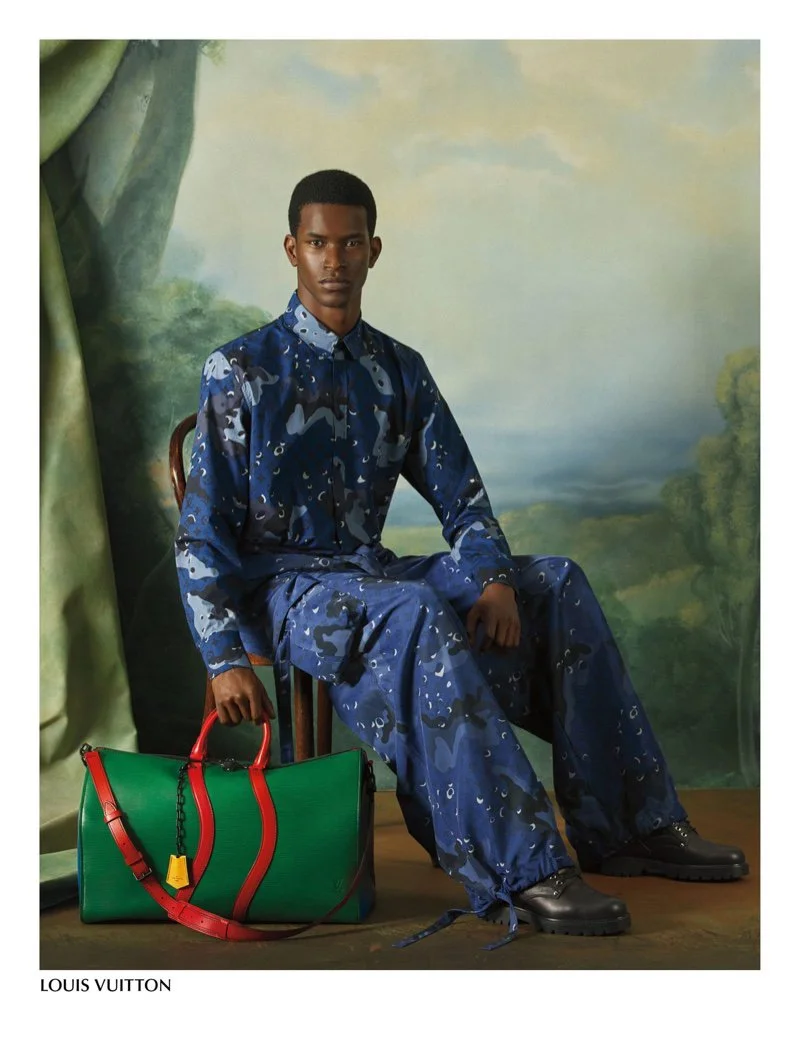 A young man sitting on a chair wearing a blue camouflage-patterned matching suit, with a large green Louis Vuitton bag with red accents placed on the floor next to him, against a painted outdoor landscape background.