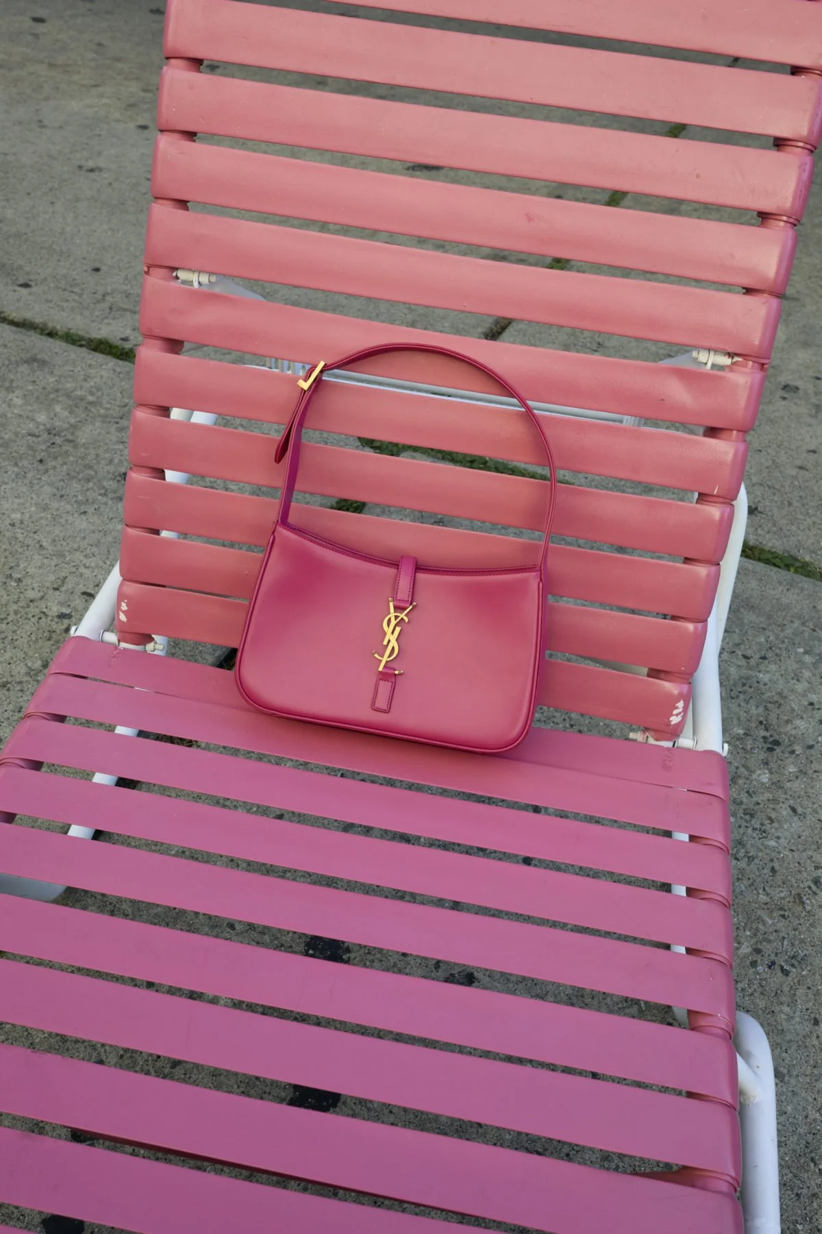 A pink handbag with a gold YSL logo is resting on a pink outdoor lounge chair with horizontal slats, outdoors on a concrete surface.