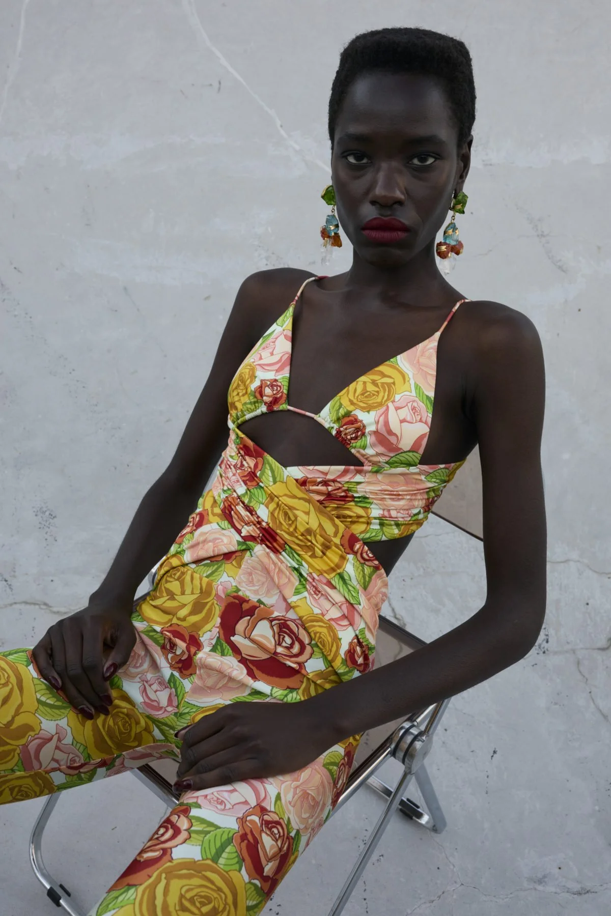 A dark-skinned woman with short hair wearing a floral dress with jumpsuit, sitting on a metal chair against a plain concrete wall, looking directly at the camera.