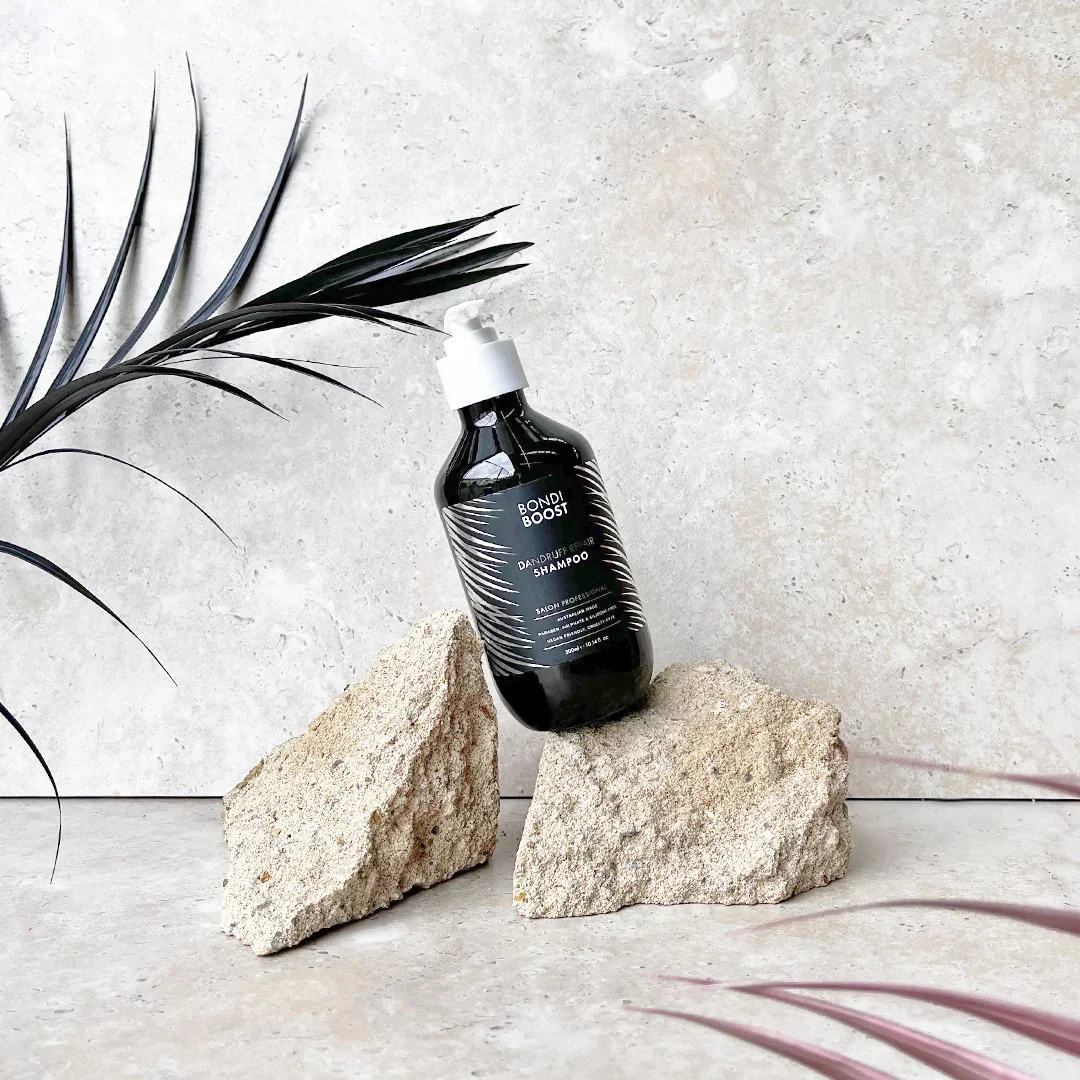 A black bottle of Bondi Boost dandruff shampoo resting on two large beige rocks against a minimalist light-colored background with a black palm leaf on the left and a pinkish plant on the right.