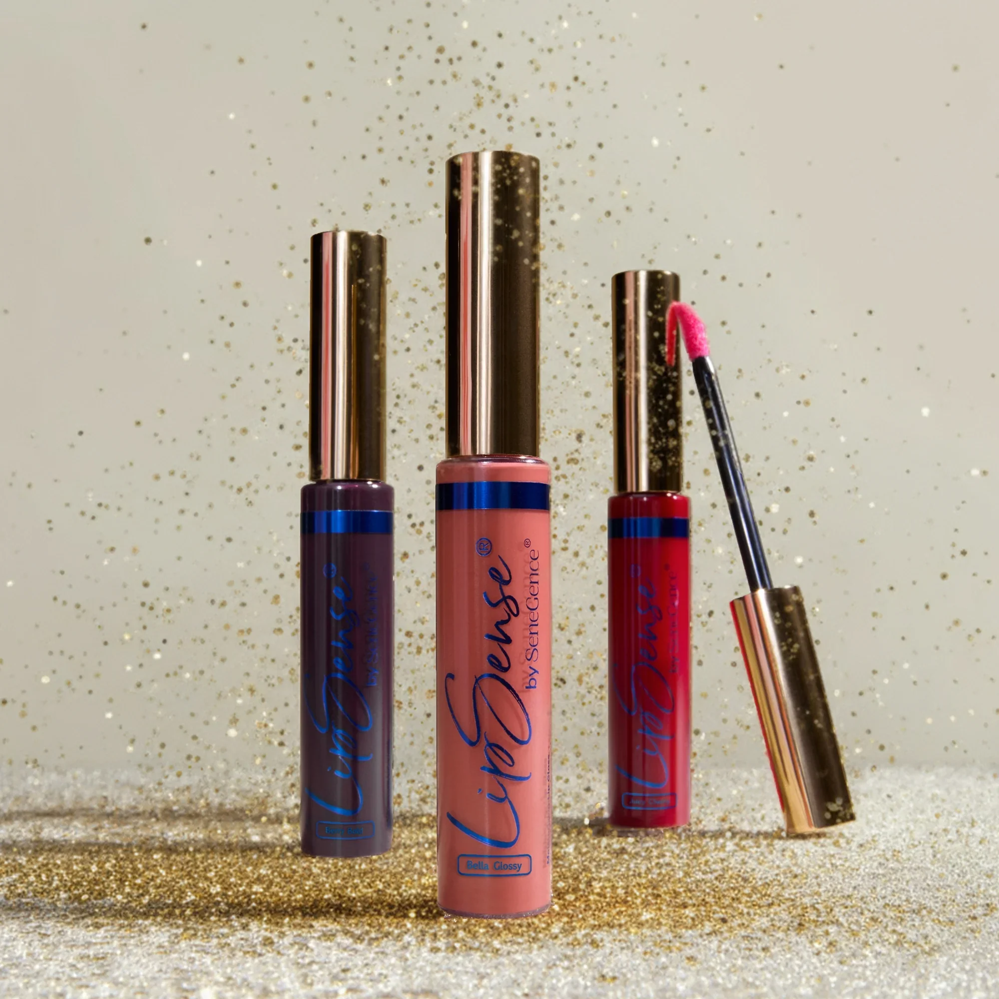 Three tubes of liquid lipstick in purple, pink, and red shades, with a gold-tipped applicator lying nearby on a gold glitter background.