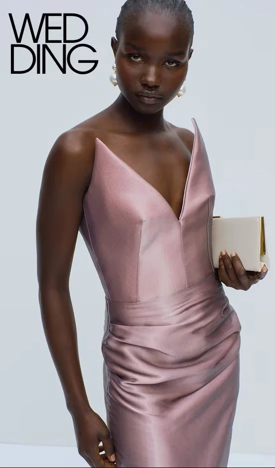 A woman with dark skin and short hair wearing a silky pink dress holding a beige clutch bag, standing against a plain light background with the text "WED DING" in the top left corner.