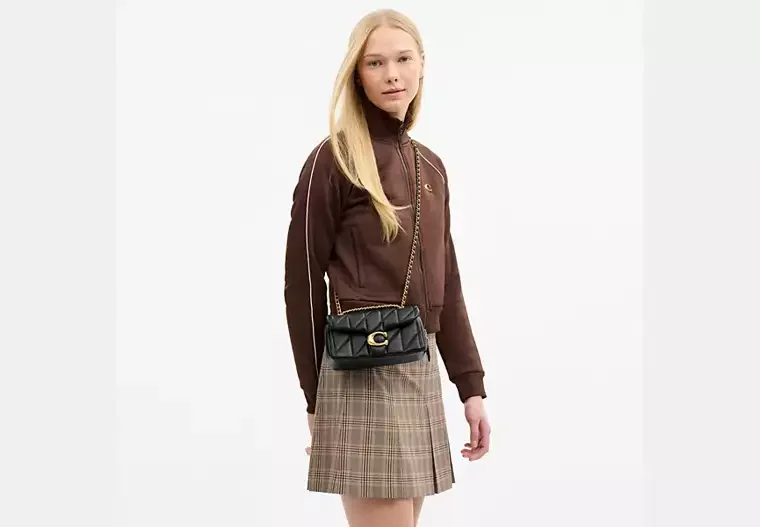 A young woman with long blonde hair wearing a brown jacket, plaid skirt, and carrying a small black purse with a gold chain strap.