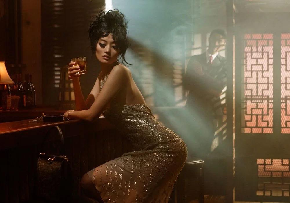 A woman with styled hair in a gold sequined dress sitting at a bar, holding a drink, with a man in the background near a patterned screen.