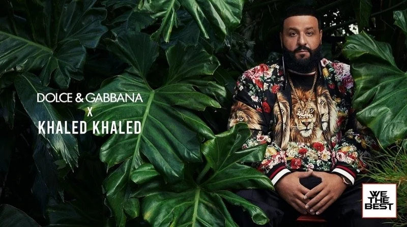 Man with a beard wearing a floral jacket with a lion face on the shirt, sitting among large green leaves, with text indicating a collaboration between Dolce & Gabbana and Khaled Khaled, and a logo saying 'WE THE BEST'.