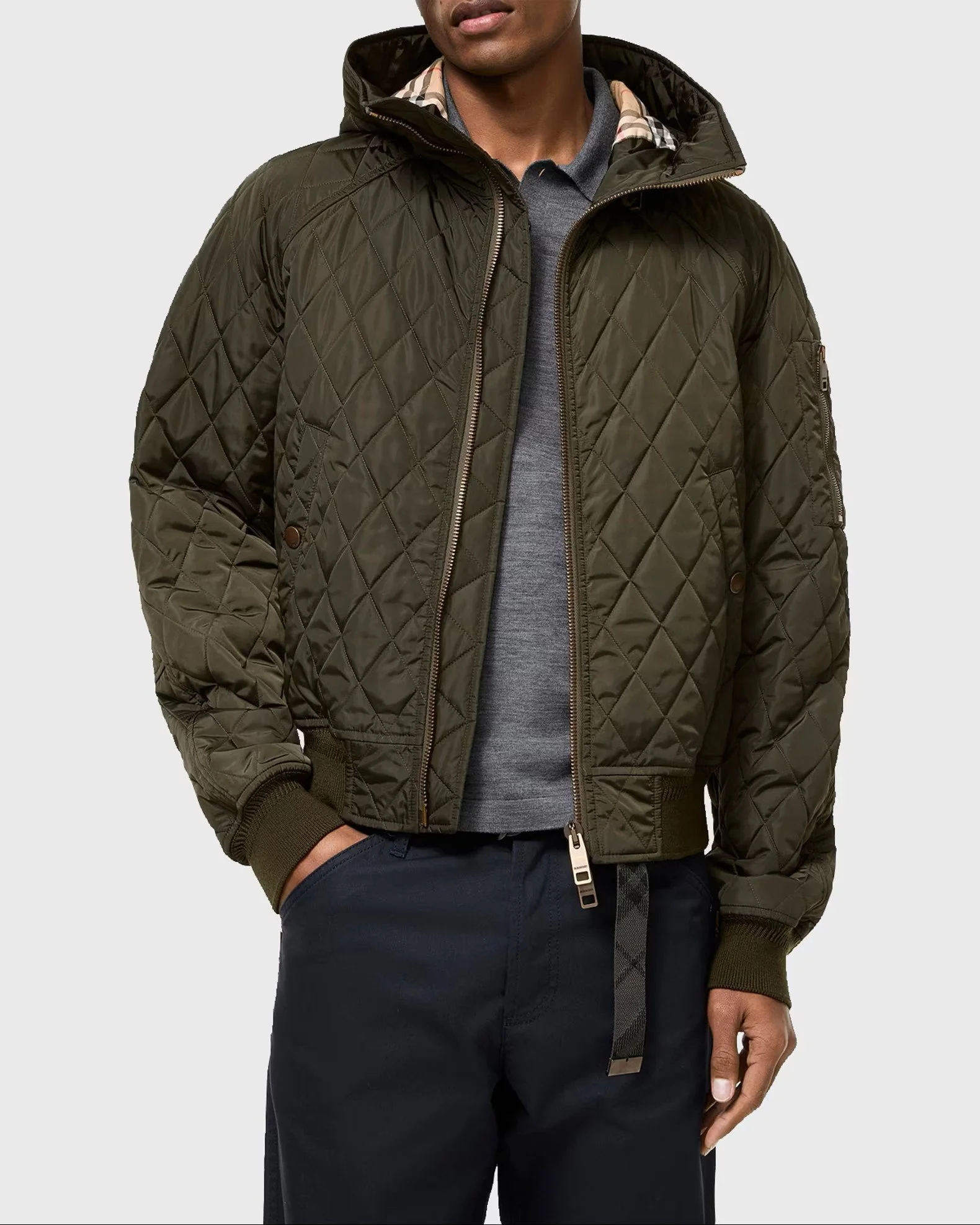 A man wearing a dark green quilted jacket with a hood, a gray shirt, and dark pants, standing with his right hand in his pant pocket.