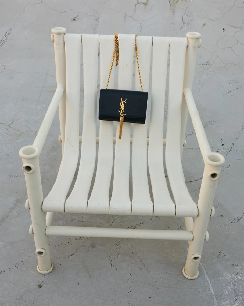 A white slatted outdoor bench with a black designer YSL clutch purse with a gold chain and tassel hanging on the backrest.