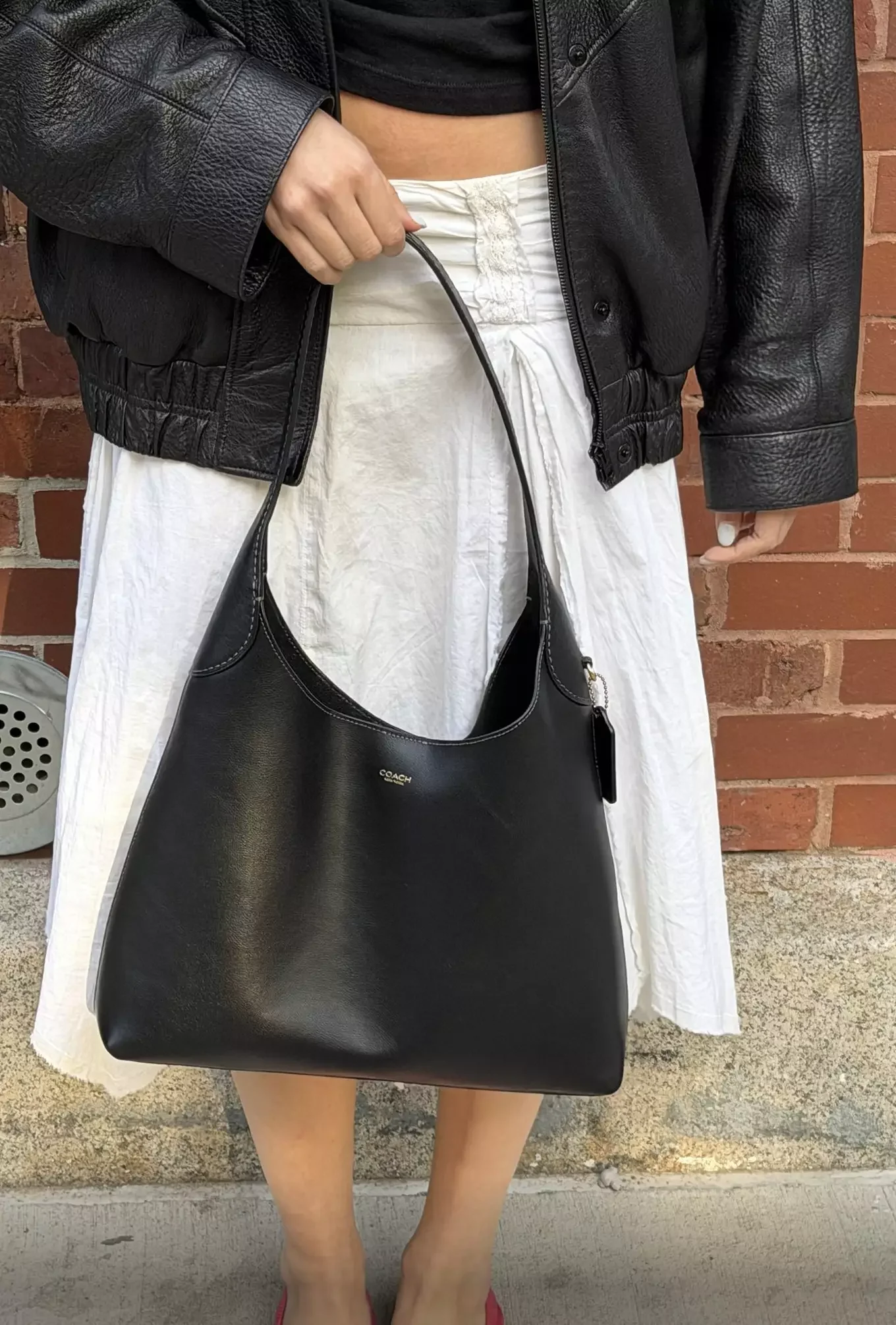 Close-up of a woman wearing a black leather jacket, white skirt, holding a black Coach handbag, standing against a brick wall.