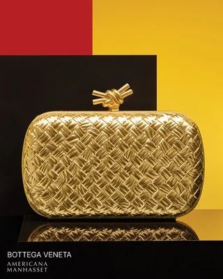 A gold-colored clutch bag with a woven pattern and a decorative bow clasp, set against a colorful background with black, red, and yellow sections.