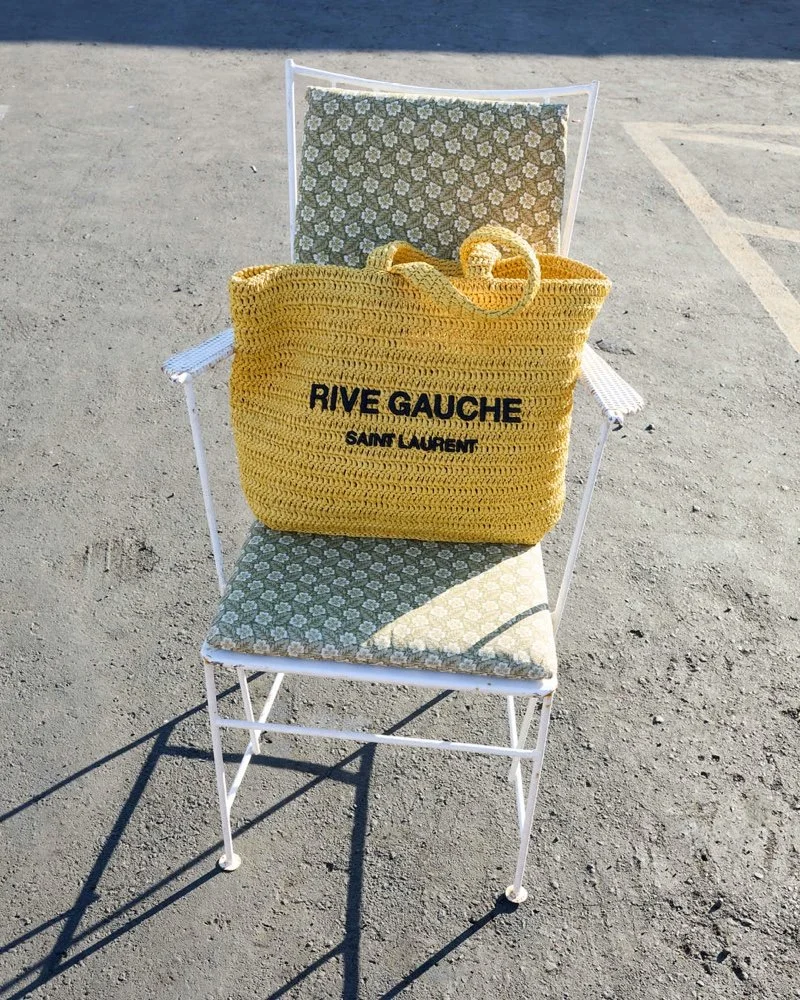 A white metal beach chair with a patterned cushion on the seat and backrest, placed on a sandy surface. A large yellow crocheted bag with black text reading 'RIVE GAUCHE SAINT LAURENT' is sitting on the chair.