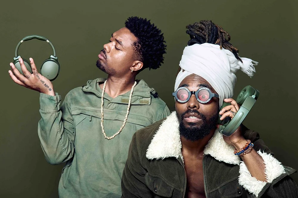 Two men with headphones, one wearing sunglasses with records on the lenses and a white headwrap, and the other holding headphones, against a green background.