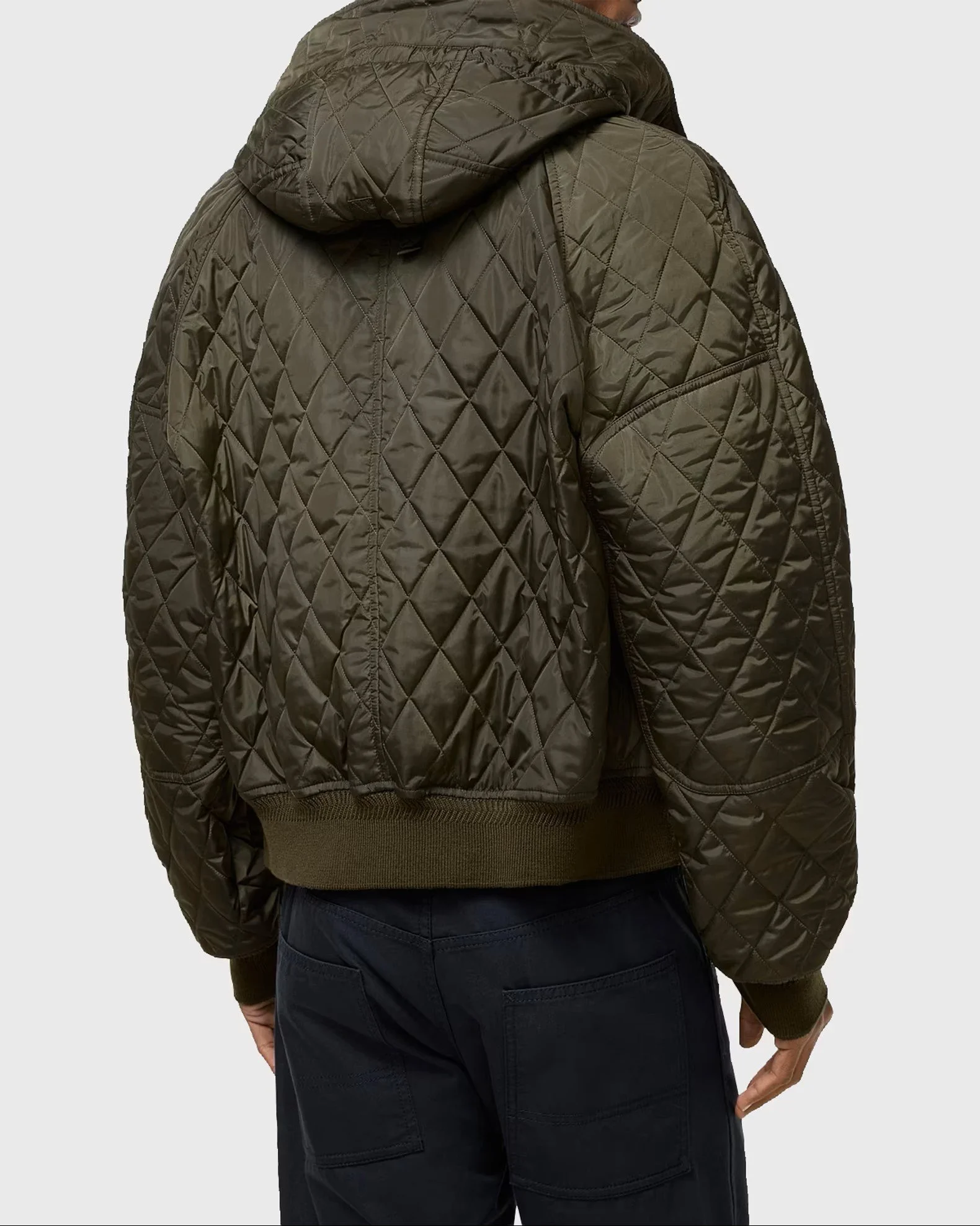 A person wearing a dark brown quilted jacket with a hood, seen from the back, with their right arm slightly bent and their hand resting near their waist, and wearing dark pants.